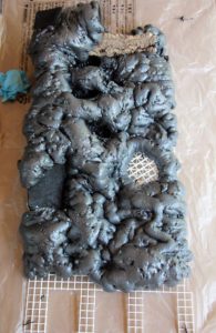 Great Stuff Pond & Stone foam is applied to the background; it will then be carved and coated with silicone which is dusted with organic materials like peat moss or coconut husk fibers to create a natural, dirt-like finish.