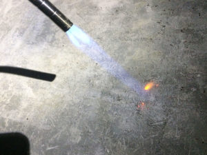 A propane hand torch burns plastic fibers from concrete surface.